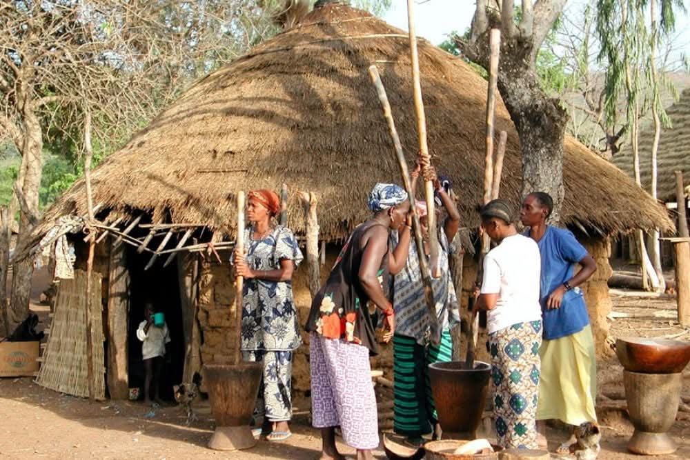 Women pounding millet outside a thatched hut